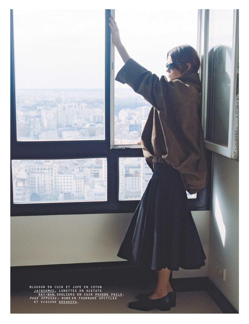 Model standing by a large window overlooking a cityscape, wearing a brown jacket and dark skirt for Harper's Bazaar France.