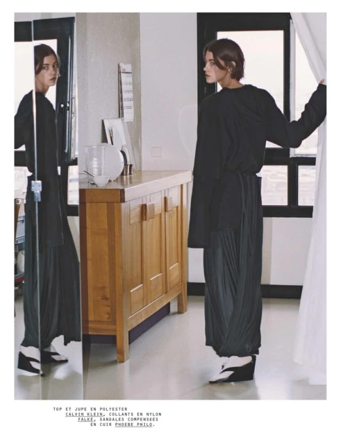 Indoor fashion shot featuring a woman in a black top and pleated skirt, accented by wedge shoes, posing near a mirror in a brightly lit room.