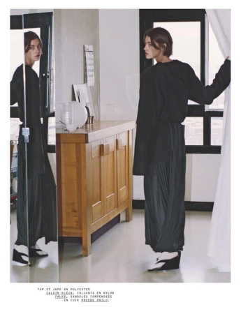 Indoor fashion shot featuring a woman in a black top and pleated skirt, accented by wedge shoes, posing near a mirror in a brightly lit room.