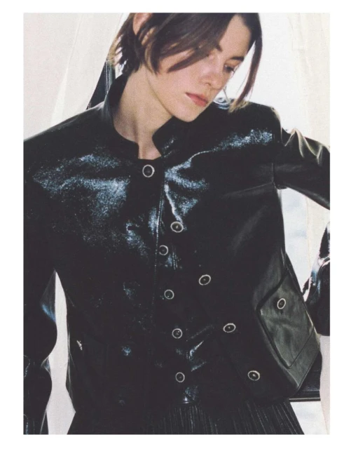 Brunette woman in black patent leather jacket poses indoors by window, showcasing high-fashion, editorial style and studio lighting.