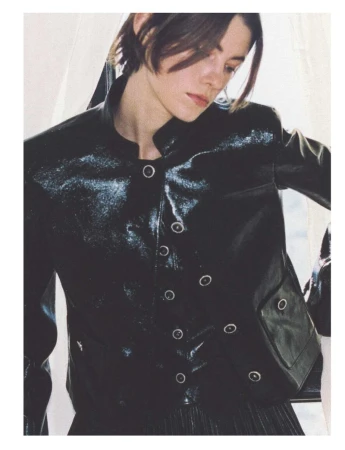 Brunette woman in black patent leather jacket poses indoors by window, showcasing high-fashion, editorial style and studio lighting.