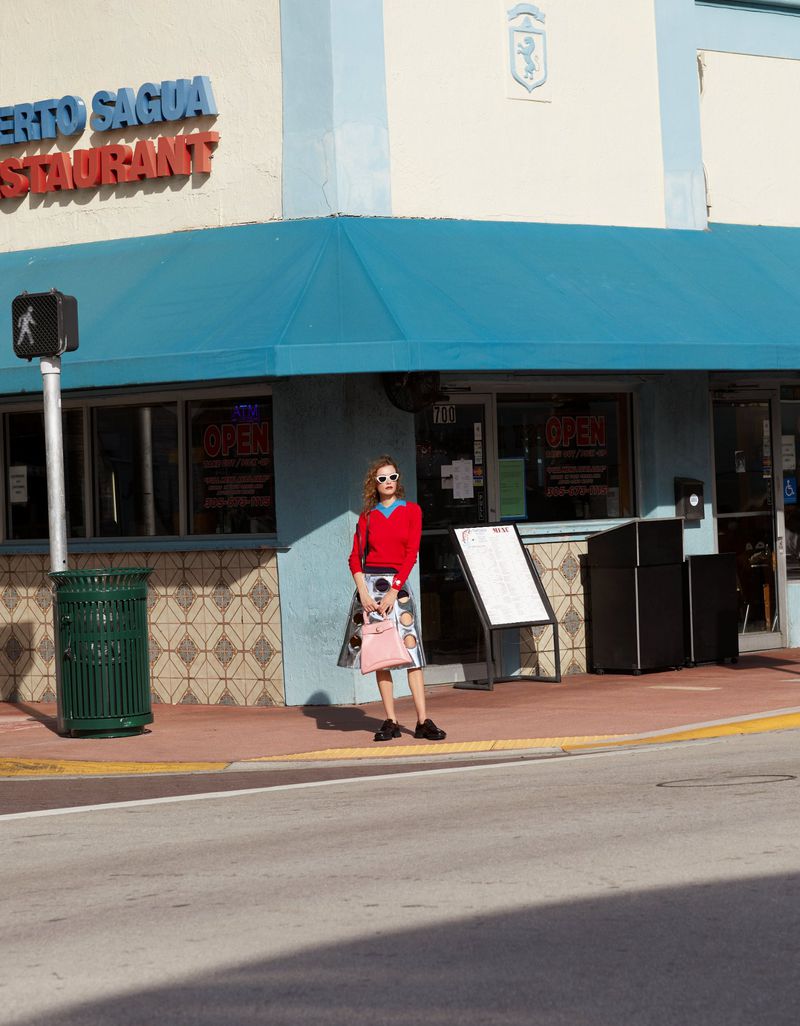 Woman in red sweater and patterned skirt stands near restaurant with pink handbag and sunglasses, captured on sunny day.
