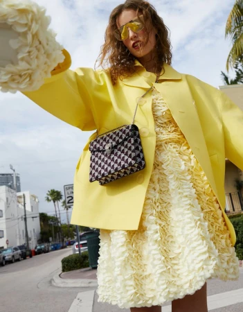 A fashionable young woman in a yellow coat and textured dress poses on a city street with palm trees, sunglasses, and a handbag.