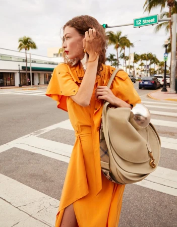Model in vibrant orange dress holding a large beige handbag on a sunny city street intersection with palm trees.