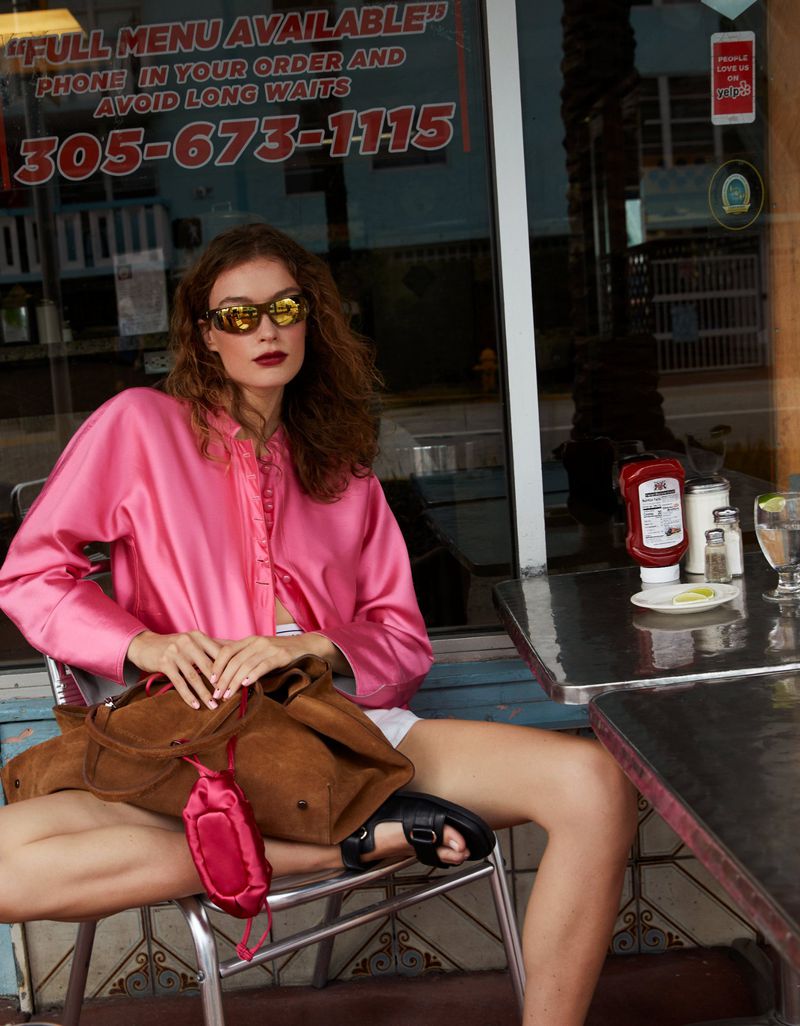 A young woman in sunglasses and a pink jacket poses at an outdoor cafe with a brown bag, next to a window with text and reflections.
