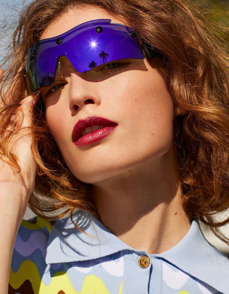 Portrait of a woman wearing futuristic visor-style sunglasses with red lipstick, a patterned shirt, and a playful, modern fashion aesthetic.