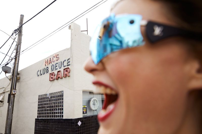 Close-up of smiling woman with sunglasses in front of vintage bar with neon sign reading MAC'S DEUCE CLUB BAR in daylight.