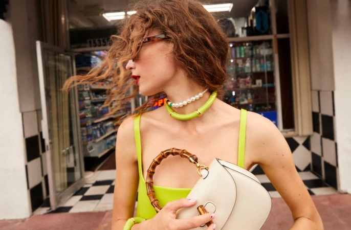 Woman in lime dress poses with white Gucci purse and pearl necklace outside store with red lipstick, sunglasses, and wavy hair.