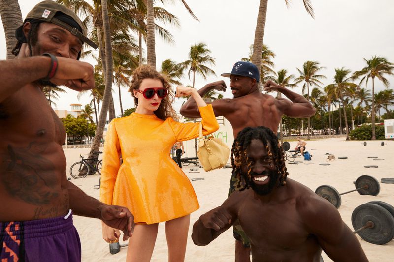 Four people pose on a beach with palm trees. A woman in an orange dress stands between three shirtless men. Weights are in the background.
