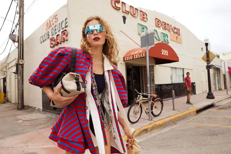 Stylish woman poses in front of Club Deuce bar with accessories, including a handbag, statement sunglasses, and jewelry in an urban setting.