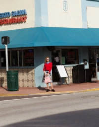 Model wearing a red sweater and patterned skirt holding a pink handbag outside a restaurant storefront
