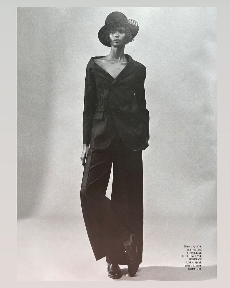 Monochrome full-length portrait of a woman in a dark blazer, wide-leg trousers, and a hat against a grey backdrop, fashion editorial style.