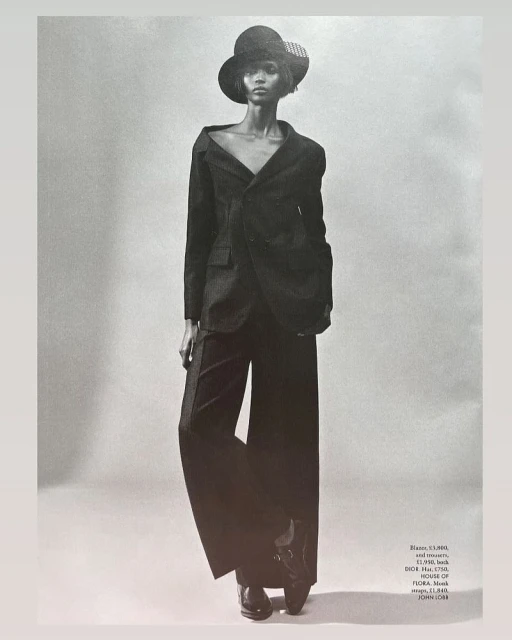 Monochrome full-length portrait of a woman in a dark blazer, wide-leg trousers, and a hat against a grey backdrop, fashion editorial style.