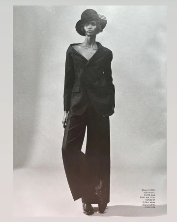 Monochrome full-length portrait of a woman in a dark blazer, wide-leg trousers, and a hat against a grey backdrop, fashion editorial style.