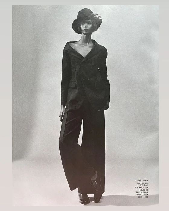 Model wearing a dark tailored suit and wide-brimmed hat against a neutral studio background in a black and white fashion photograph.