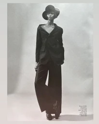 Model wearing a dark tailored suit and wide-brimmed hat against a neutral studio background in a black and white fashion photograph.