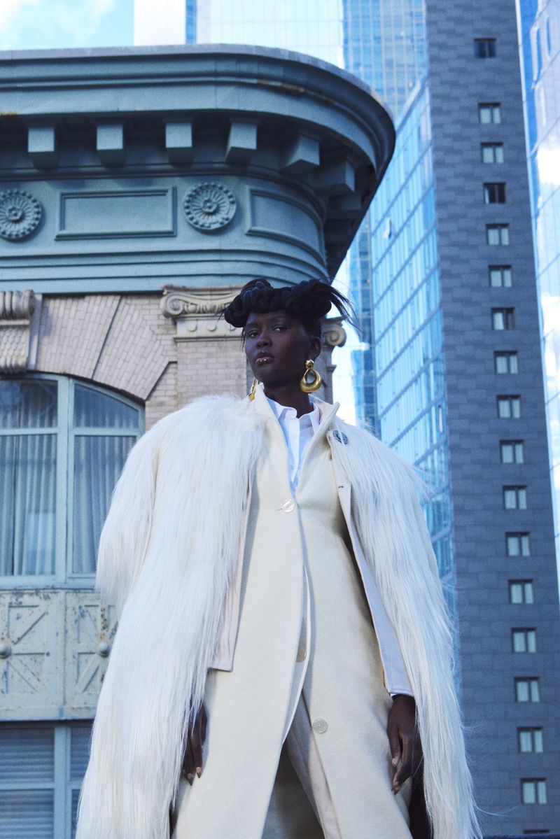 Elegant adult female in cream coat poses against cityscape background, featuring modern buildings and architectural details, fashion editorial.