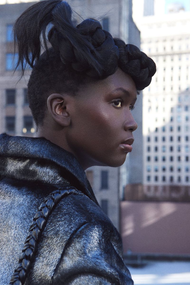 Profile shot of a dark-skinned woman with intricate braided updo wearing a dark leather jacket with braided detailing, against a city backdrop.