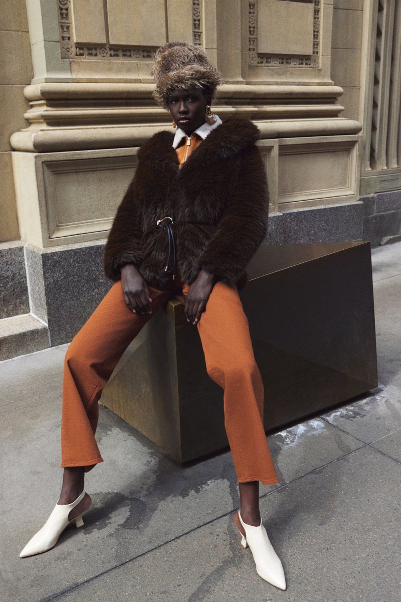 A single Black female model in an earthy-toned fashion shoot poses on a cube outdoors, wearing a fur jacket, tan pants, and white slingback heels.