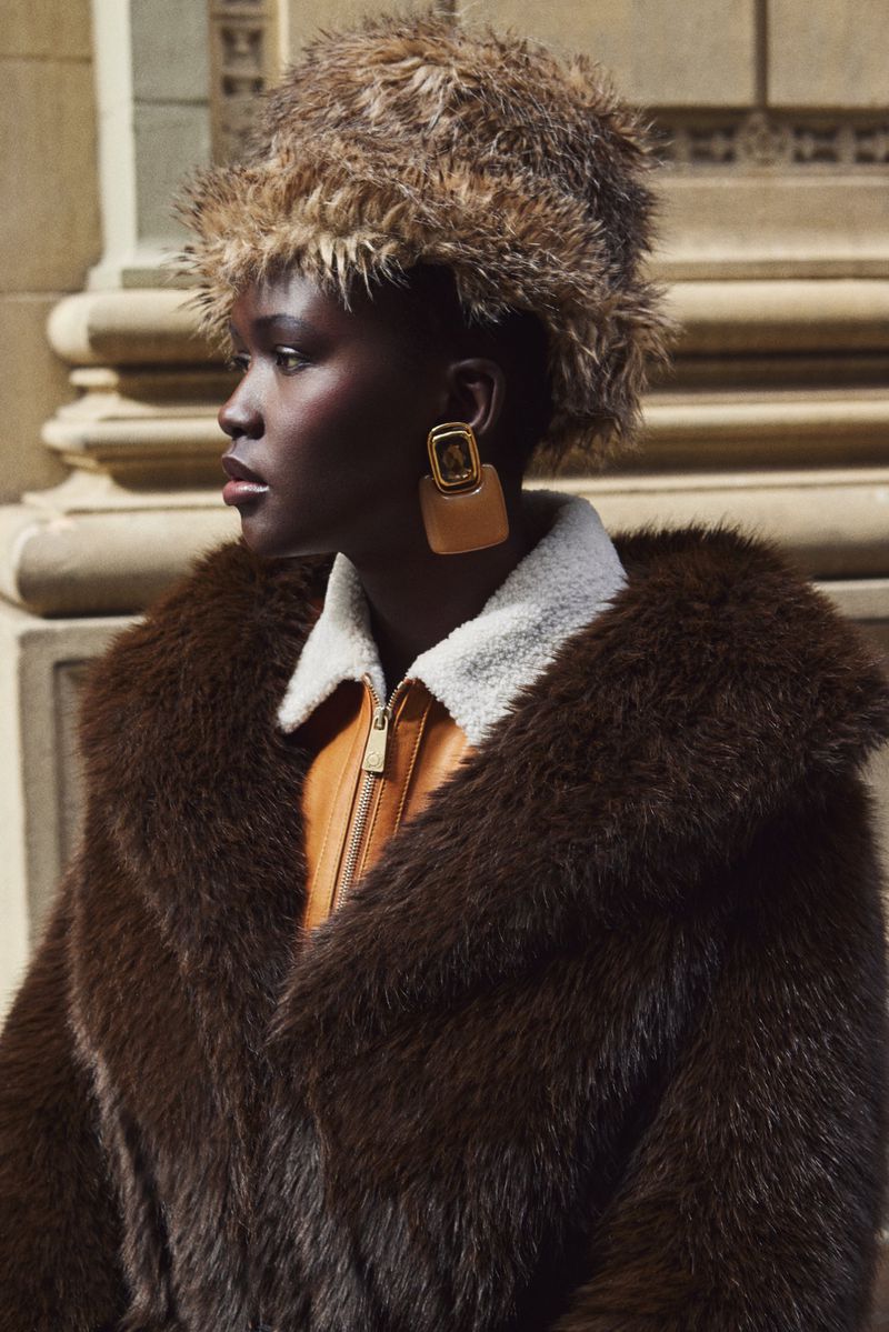 Dark-skinned woman in fur hat and coat, accessorized with tan earrings, poses in front of a sandstone building with architectural details.