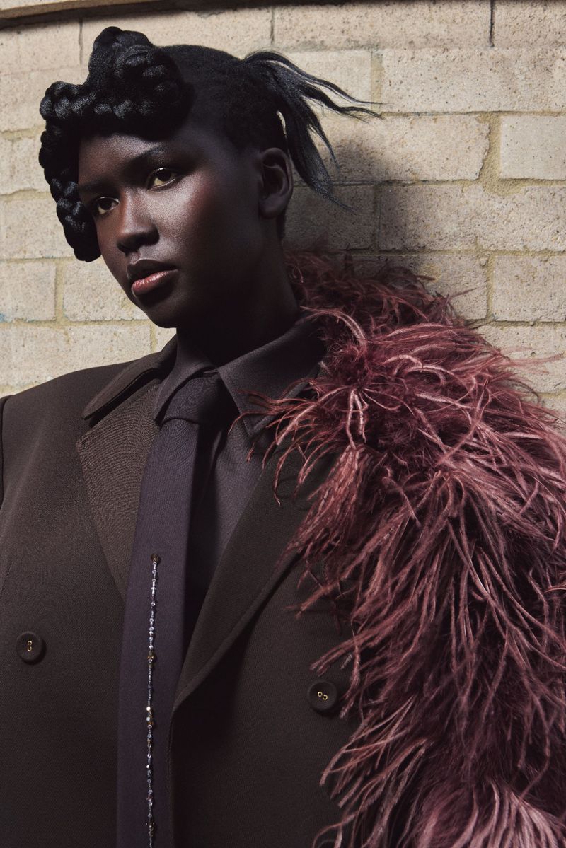 Portrait of African American woman in a dark brown suit with a feather boa in front of a brick wall in a studio.