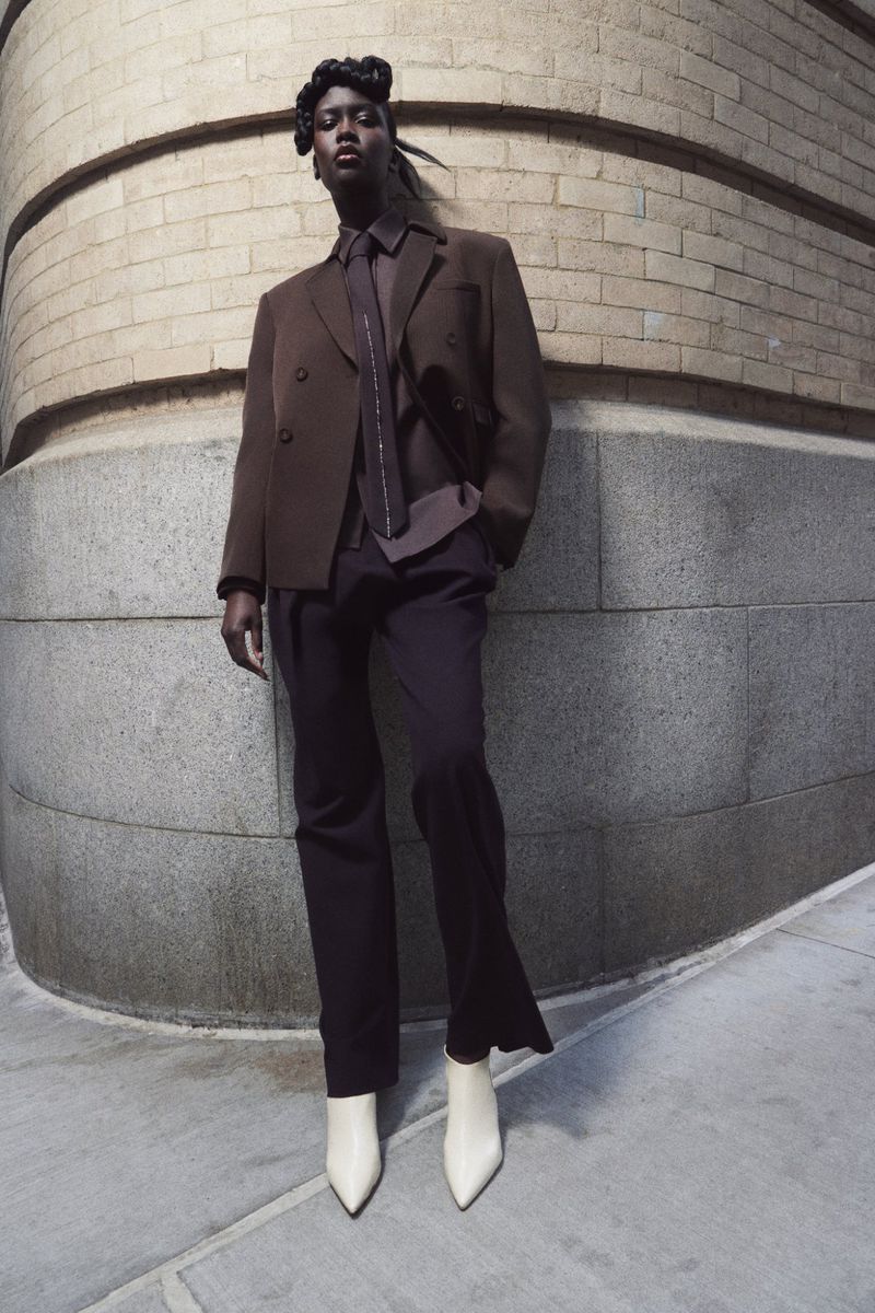A Black female model strikes a pose outdoors, wearing a brown blazer and trousers paired with white boots, set against a textured wall.