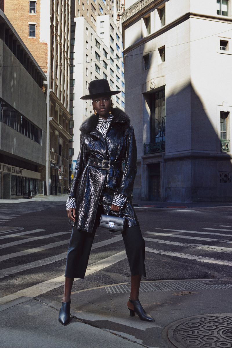 Fashionable woman in a black sequin coat and hat posing on a city street with buildings in the background, wearing ankle boots.