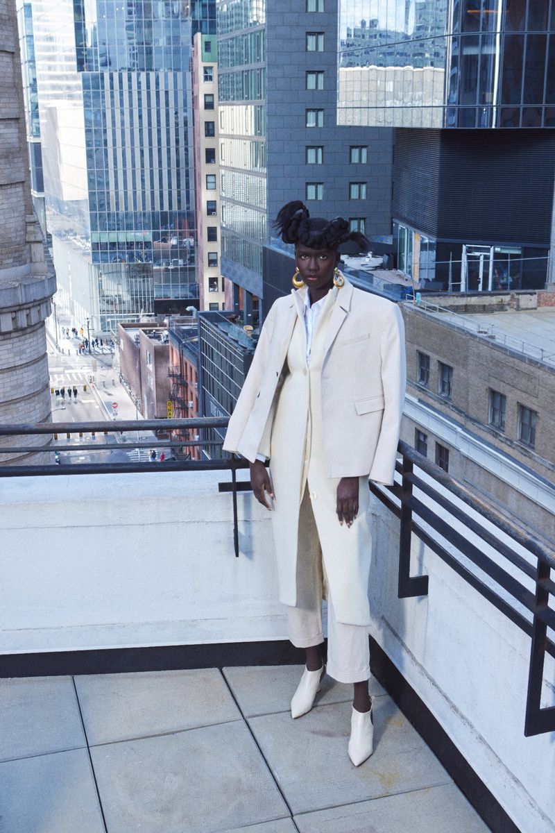 Stylish woman in a cream-colored suit and white boots poses on a rooftop with a city skyline view, capturing a fashion-forward moment.