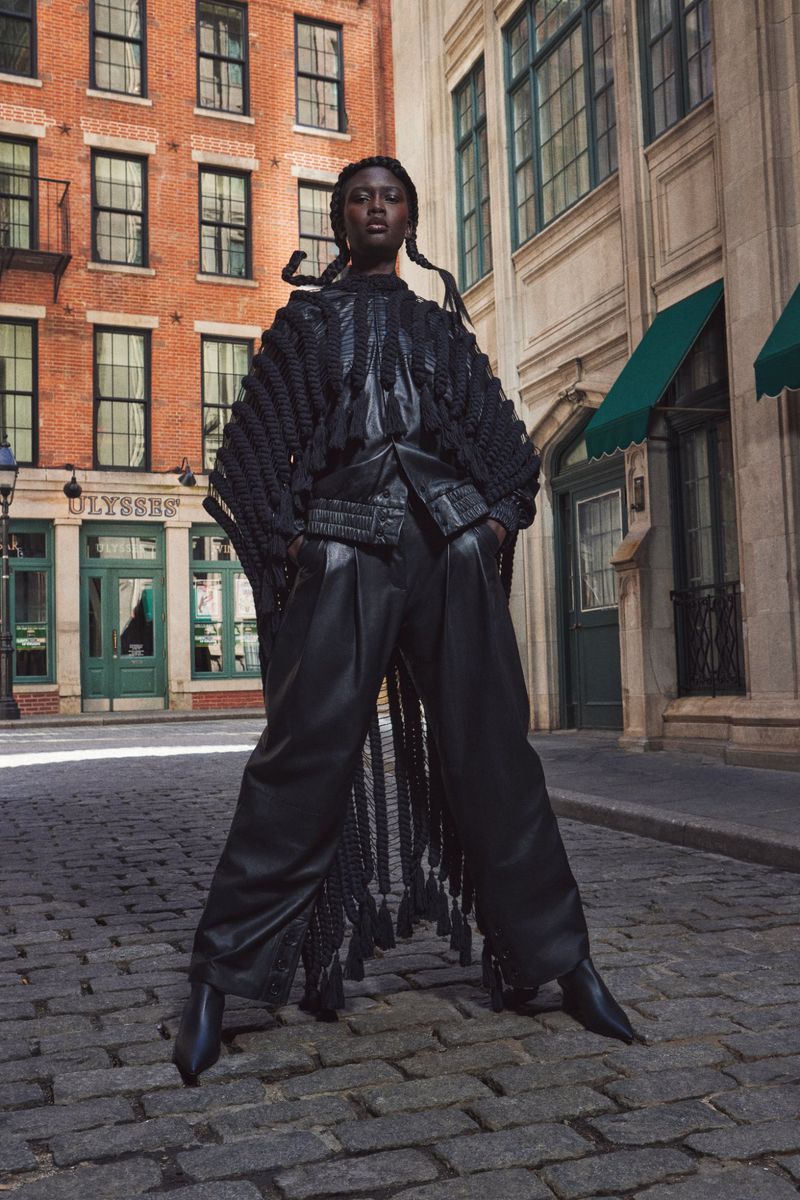 Dark-skinned model poses on cobblestone street in black poncho, leather pants and boots with vintage buildings in the background.