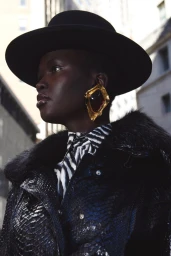 Model wearing a black wide-brim hat, statement gold earring, zebra-print shirt, and dark textured jacket against an outdoor street backdrop.