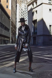 Model wearing a black hat, patterned coat, and dark trousers standing on a city street between tall buildings.