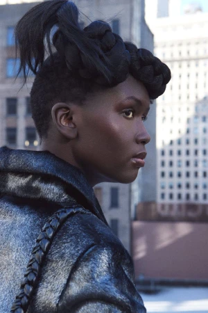Model Awuor Dit in profile with elaborate braided hairstyle wearing a dark textured coat against an urban building exterior.