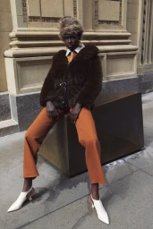 Model Awuor Dit in brown fur jacket and rust trousers sitting on a block before a stone architectural building exterior