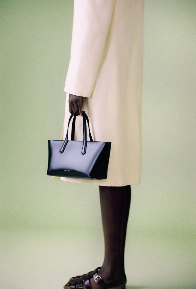 Fashion editorial shot: Black leather handbag held by a light-skinned model in a tan trench coat, paired with sandals. Studio setting.