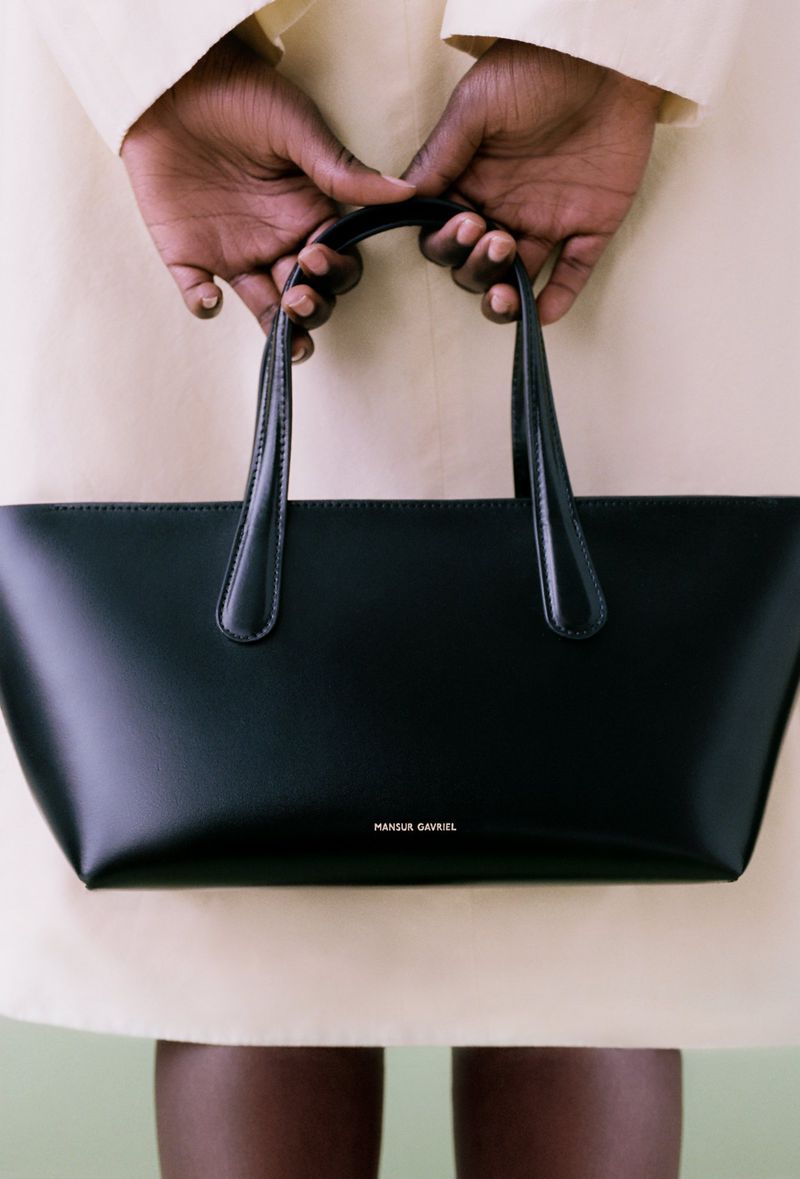 A black leather Mansur Gavriel tote bag is held by a dark-skinned person wearing a light-colored jacket against a pale green background.