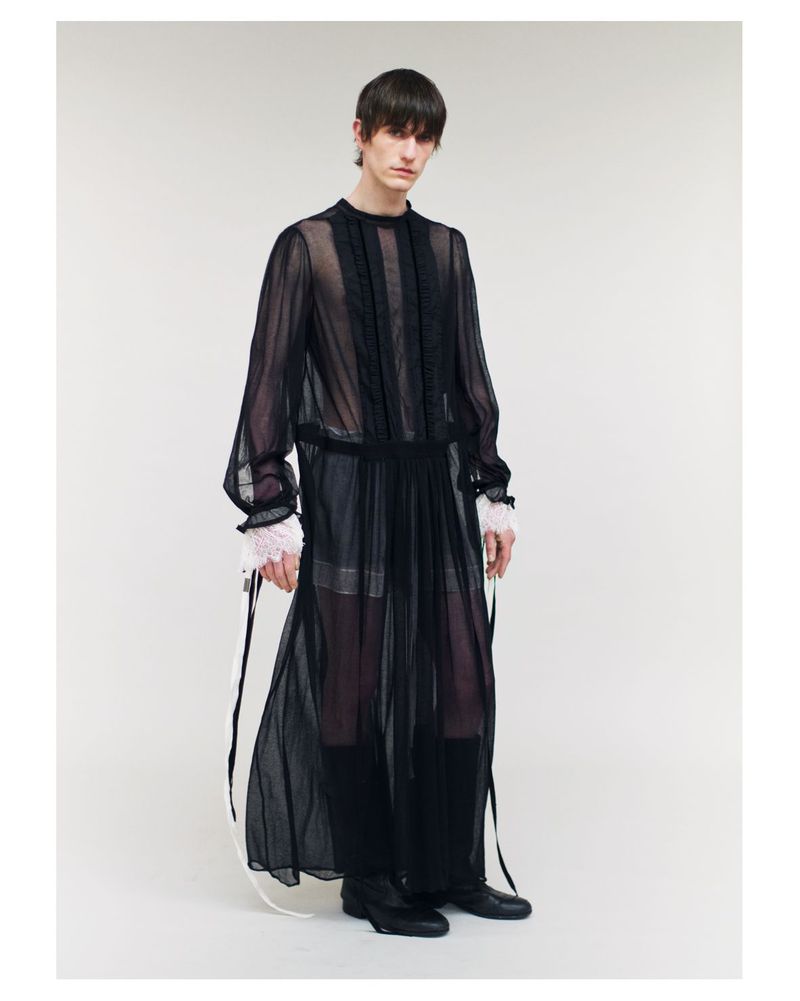 Full-length studio shot of a slender male fashion model wearing a sheer black, ankle-length gown with an opaque underlayer. 