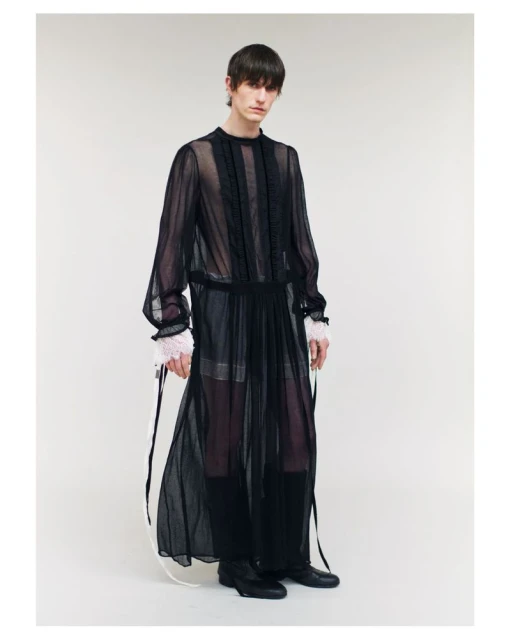 Full-length studio shot of a slender male fashion model wearing a sheer black, ankle-length gown with an opaque underlayer. 