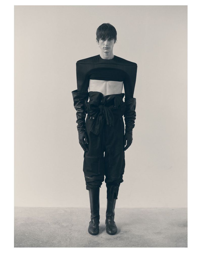 Model wearing sculptural black structured top and high leather boots standing against a neutral studio backdrop