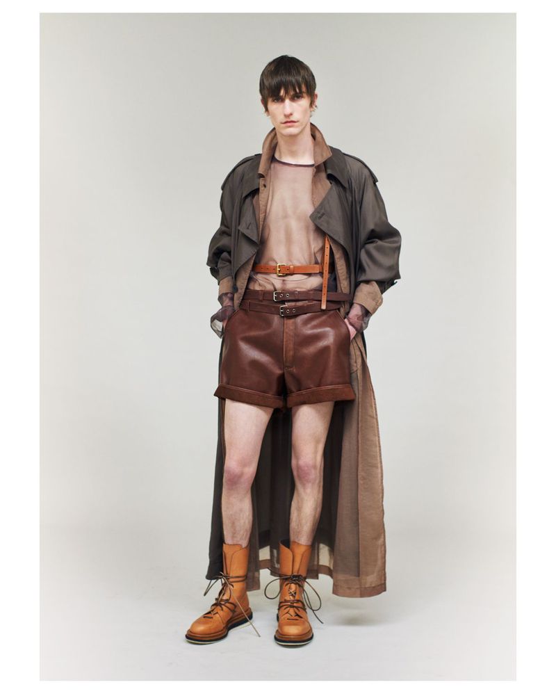 Fashion editorial shot featuring a male model in a brown trench coat, brown shorts, a beige top, and brown boots, posing in a studio.