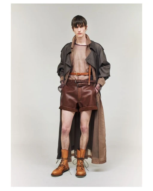 Fashion editorial shot featuring a male model in a brown trench coat, brown shorts, a beige top, and brown boots, posing in a studio.