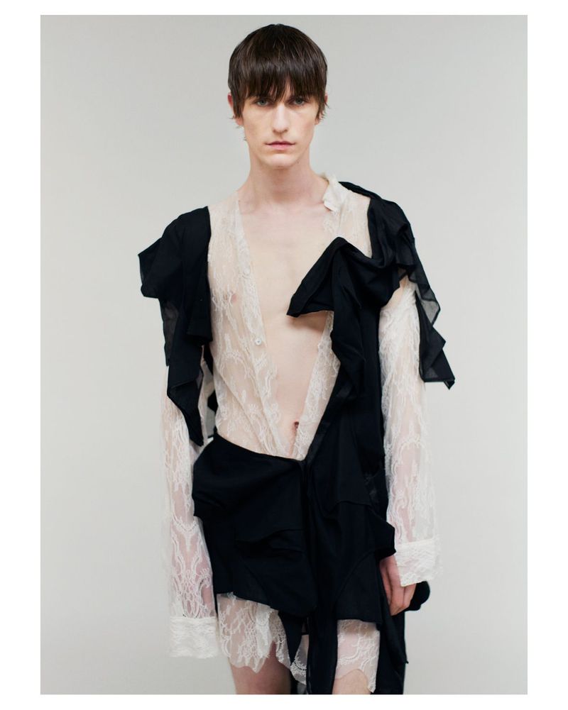 A slender, fair-skinned male model poses in a runway style with dark hair, wearing a lace top with long sleeves and black fabric accents.