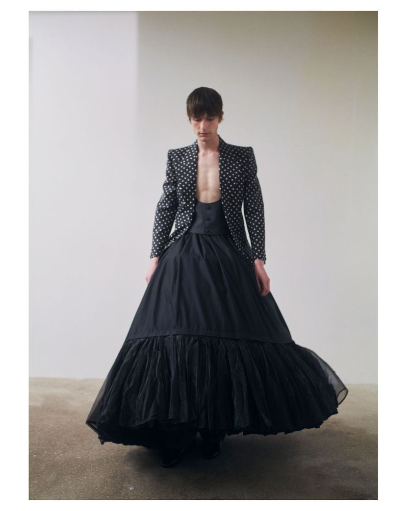 A person poses in a polka dot jacket and black skirt in an indoor photo studio, featuring dramatic fashion and a light, airy backdrop.