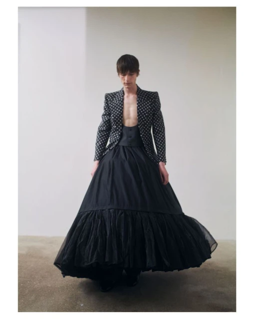A person poses in a polka dot jacket and black skirt in an indoor photo studio, featuring dramatic fashion and a light, airy backdrop.