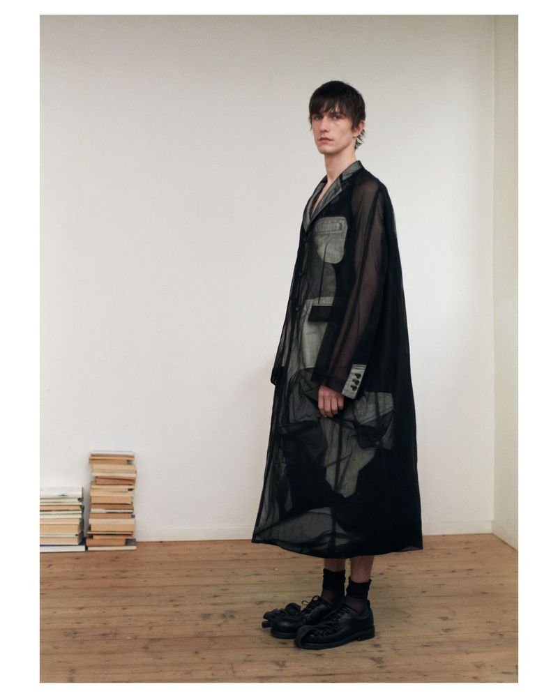 Male fashion model poses in a semi-sheer black trench coat, black dress shoes, and dark socks indoors, stacked books in the background.