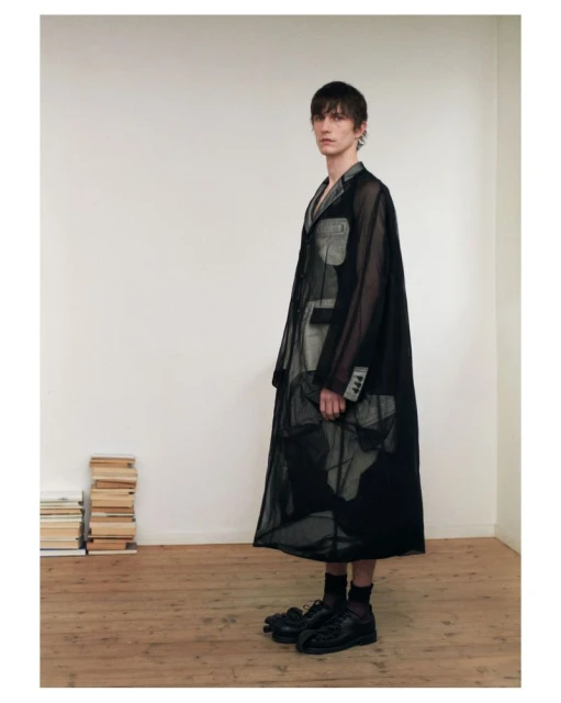 Male fashion model poses in a semi-sheer black trench coat, black dress shoes, and dark socks indoors, stacked books in the background.