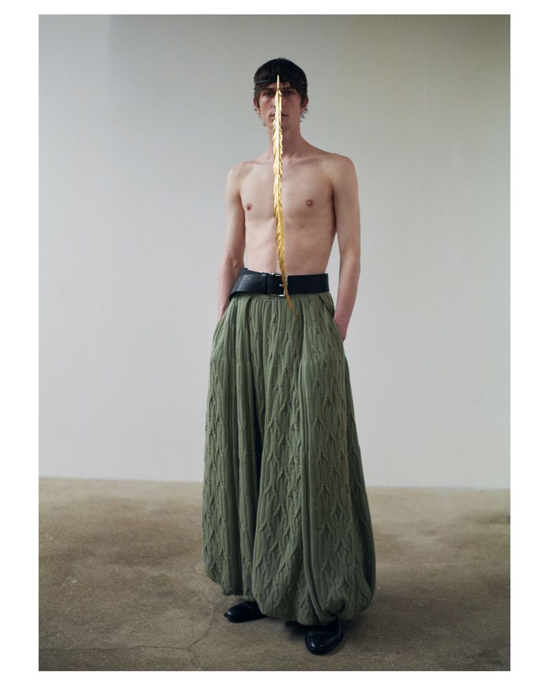 Fashion portrait of a shirtless male model wearing wide-legged green trousers and a gold forehead prop against a white wall.