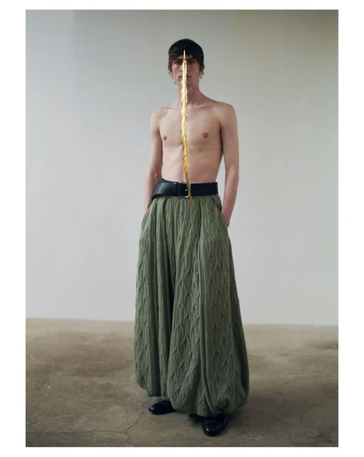 Fashion portrait of a shirtless male model wearing wide-legged green trousers and a gold forehead prop against a white wall.