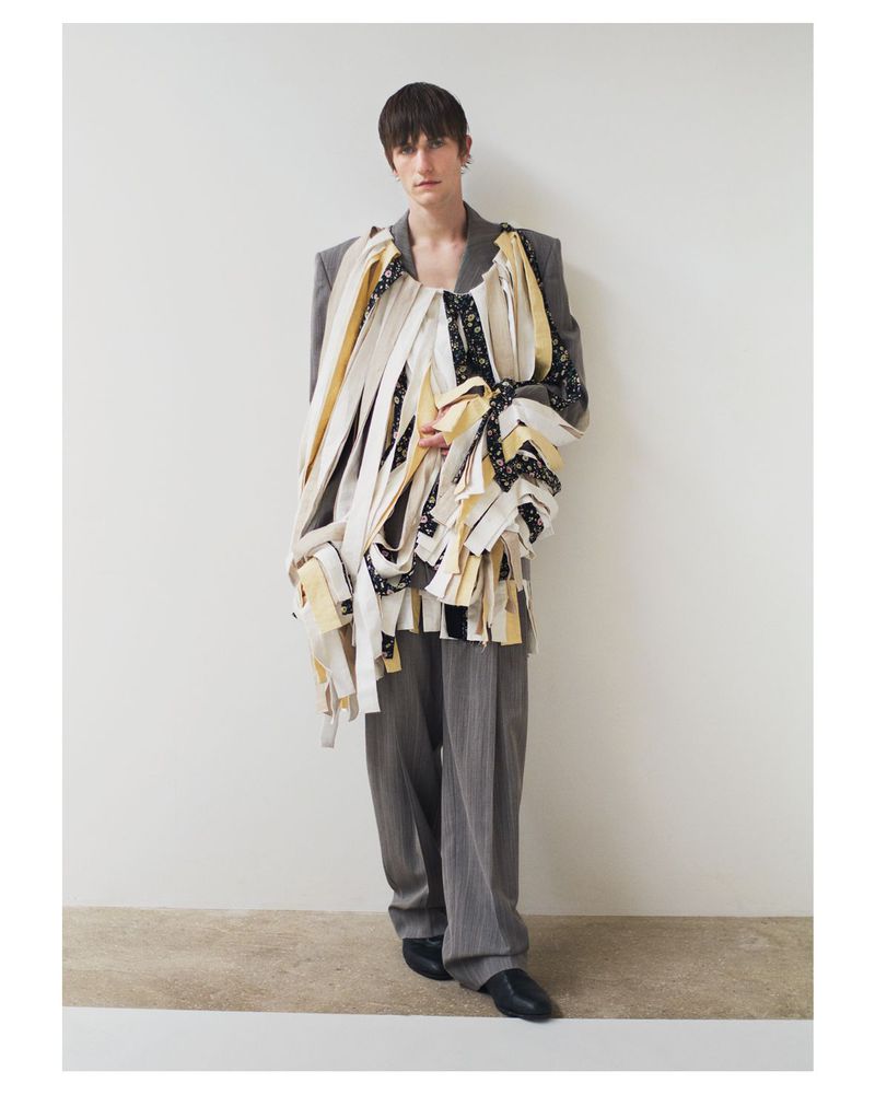 Full-length shot of adult male fashion model in gray suit, draped in multicolor patterned fabric, posing against white wall backdrop in photo studio.