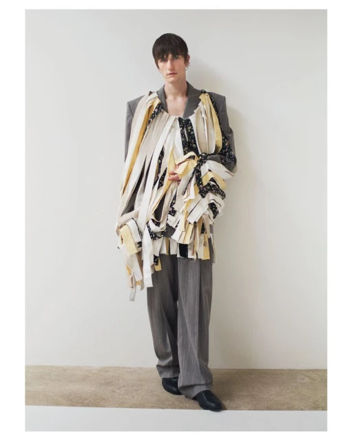 Full-length shot of adult male fashion model in gray suit, draped in multicolor patterned fabric, posing against white wall backdrop in photo studio.