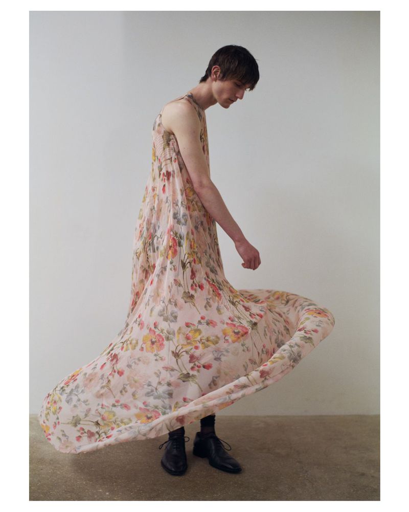 A slender man in a flowing floral dress stands against a pale backdrop, showcasing the dress's delicate pattern and elegant drape.